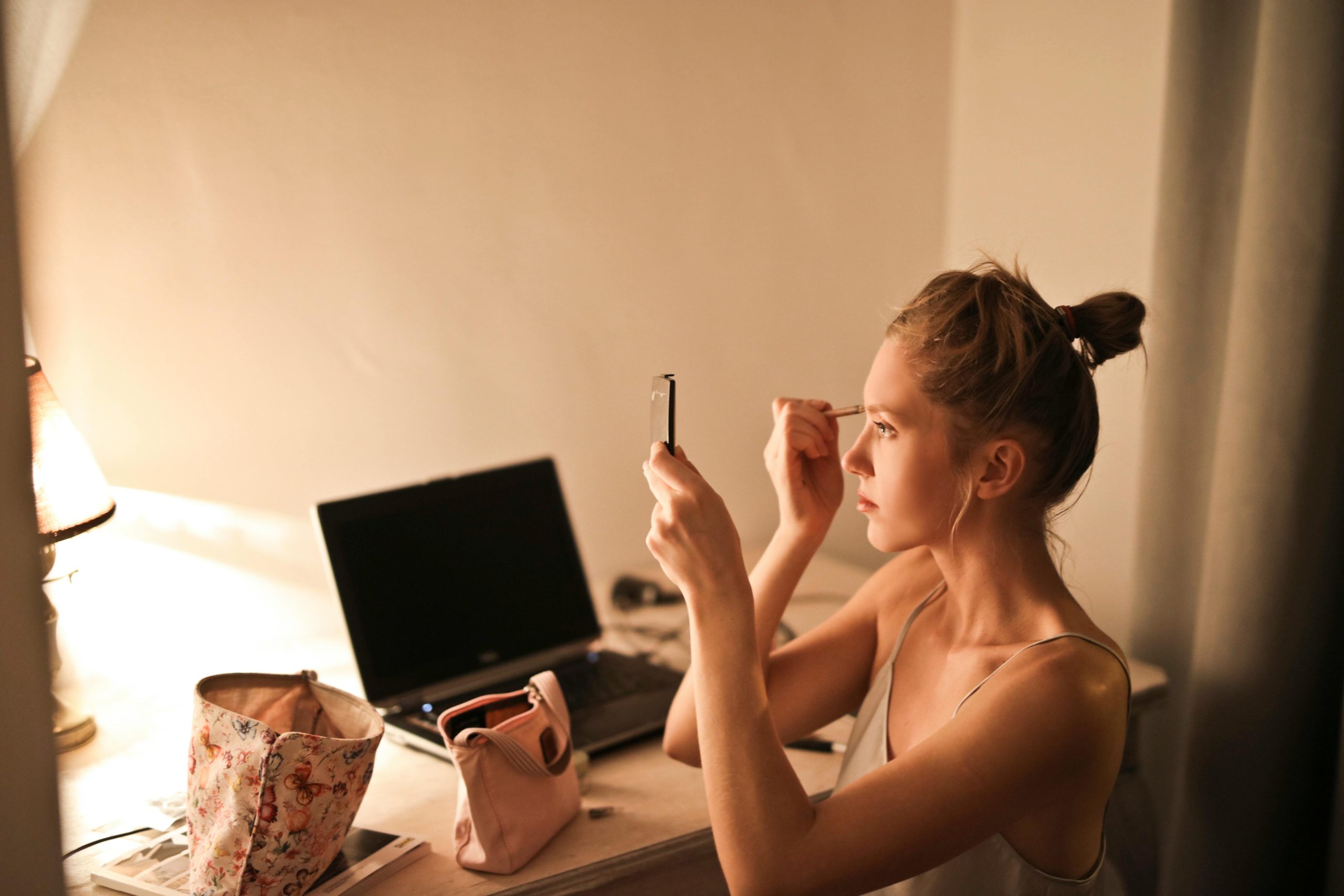 A young woman applies makeup at her vanity table, illuminated by soft light, in a cozy home setting.