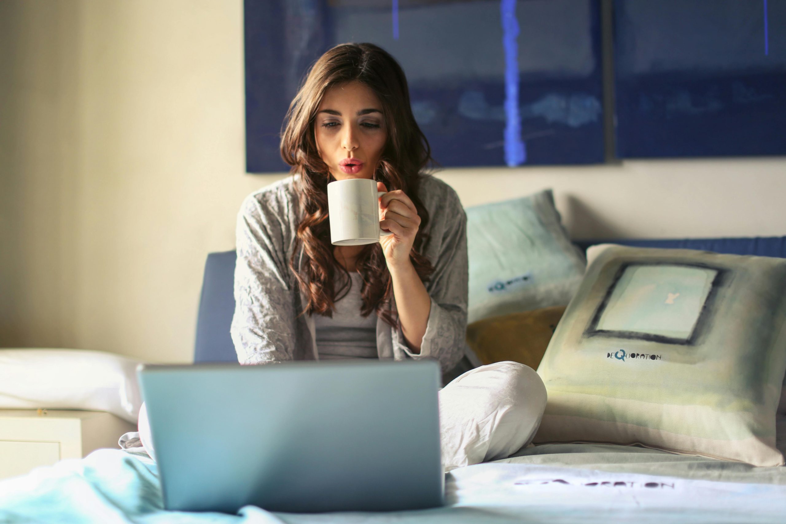 A woman enjoying coffee while working from home in a cozy bedroom setting with a laptop.